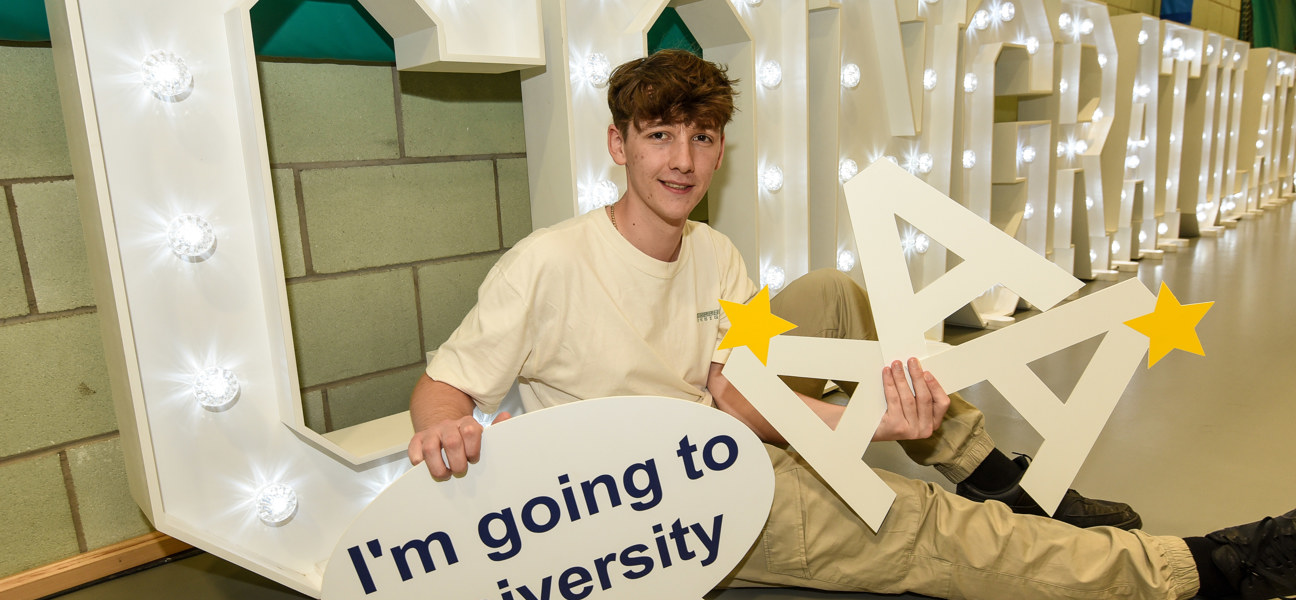 student sat holding up university sign in front of congratulations letters